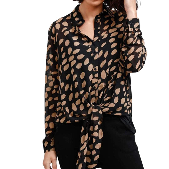A woman wearing a black long-sleeve button-up blouse with a beige abstract spotted pattern, tied at the waist, paired with black pants against a white background.