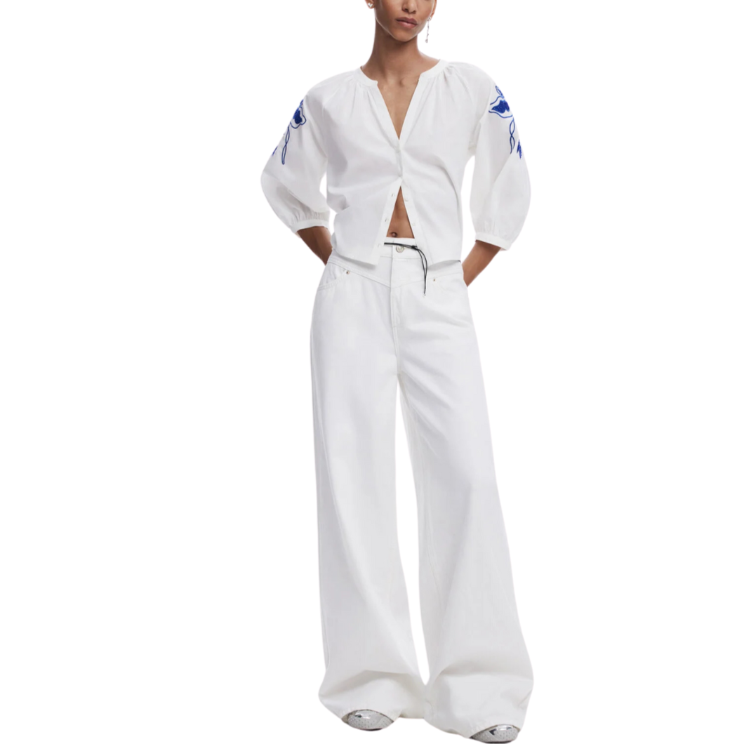 A female model wearing a white wide leg jeans with a button and zipper closure, and matching white top with sleeve details.