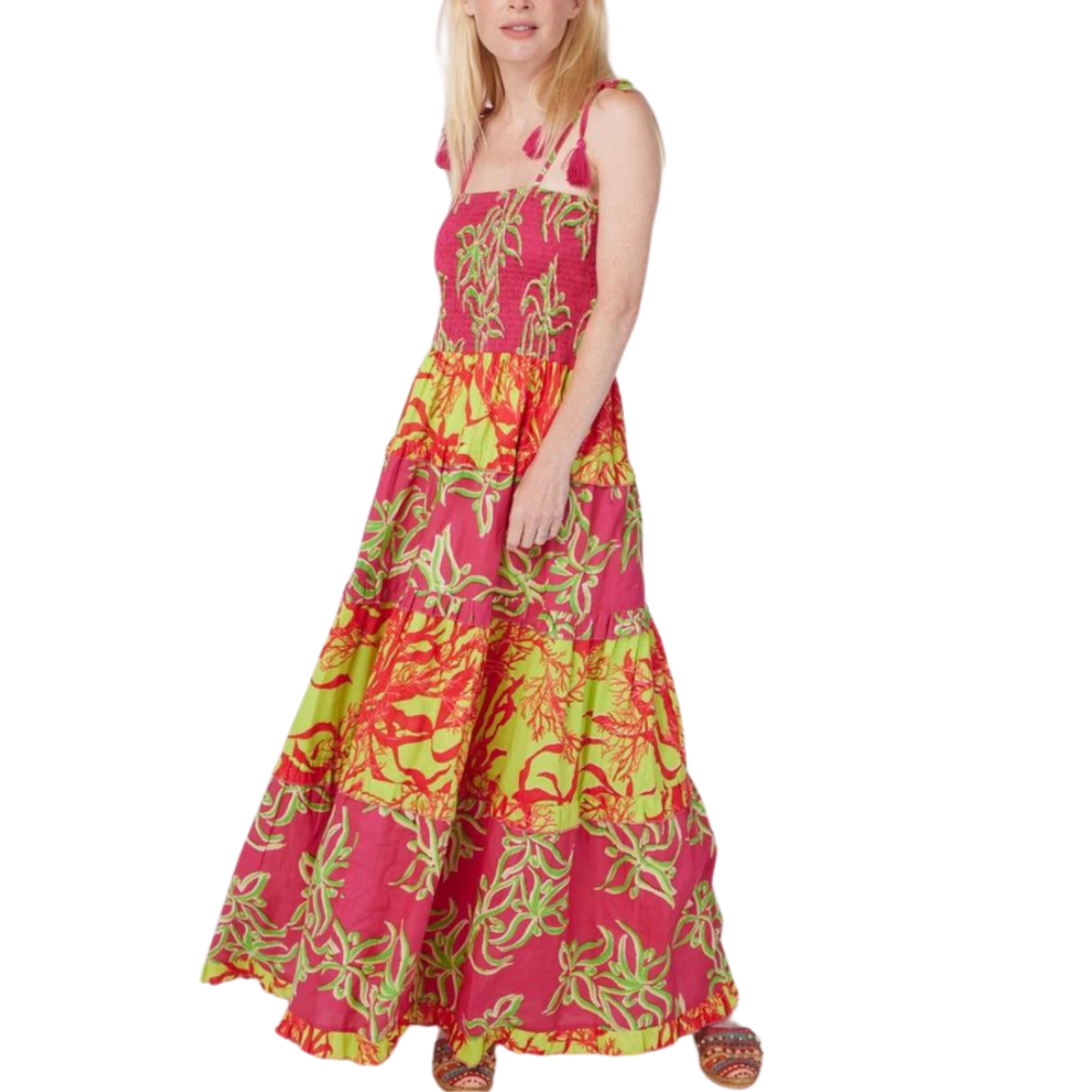 A woman wearing a floral maxi dress with a tropical print in pink, green, and yellow hues. The dress features a smocked waist and a tassel detail.