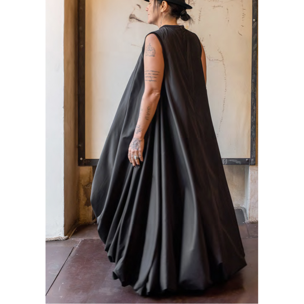 A model wearing a long, black, double-layered dress with a dramatic drape and ribbed trim, featuring a puff-ball hem and inset pockets on the seams.