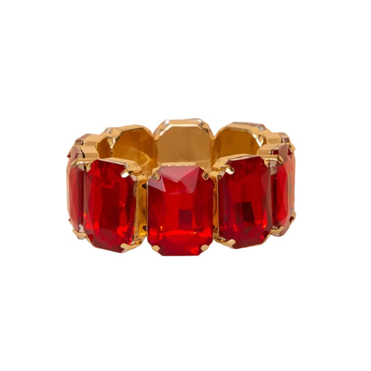 A gold-plated wide cuff bracelet featuring five large, faceted red rectangular crystals, displayed on a white pedestal against a warm beige background.