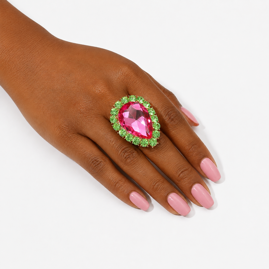 A close-up of a hand with pink manicured nails wearing a large teardrop-shaped pink gemstone ring surrounded by a halo of small green stones, set against a plain white background.