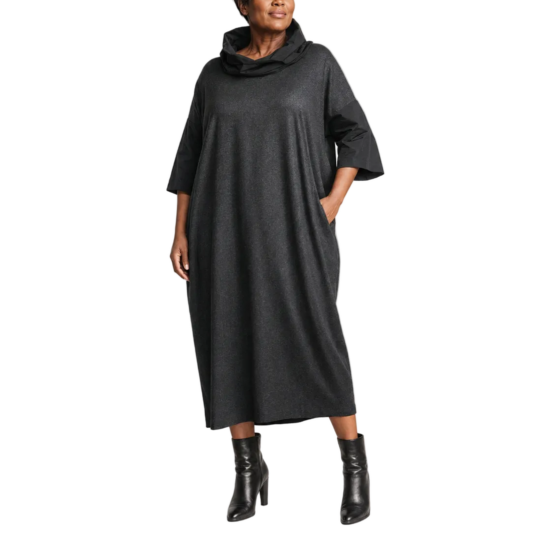 A woman modeling a dark charcoal gray cowl-neck maxi dress with elbow-length sleeves and side pockets, paired with black ankle boots, against a solid light beige studio background.