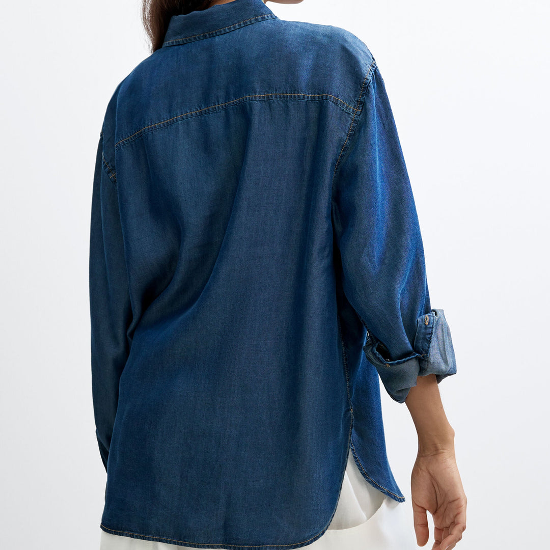 A woman shown from the back wearing a dark blue denim shirt with rolled-up sleeves and white pants, photographed against a plain light gray studio background.