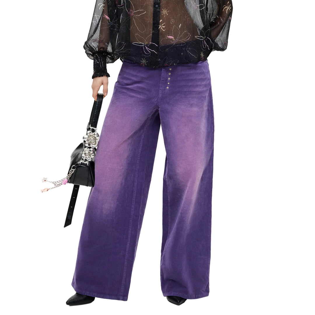 A model stands against a light gray studio background wearing a sheer black long-sleeve button-up shirt with a subtle celestial print, paired with wide-leg purple gradient pants and holding a small black handbag.