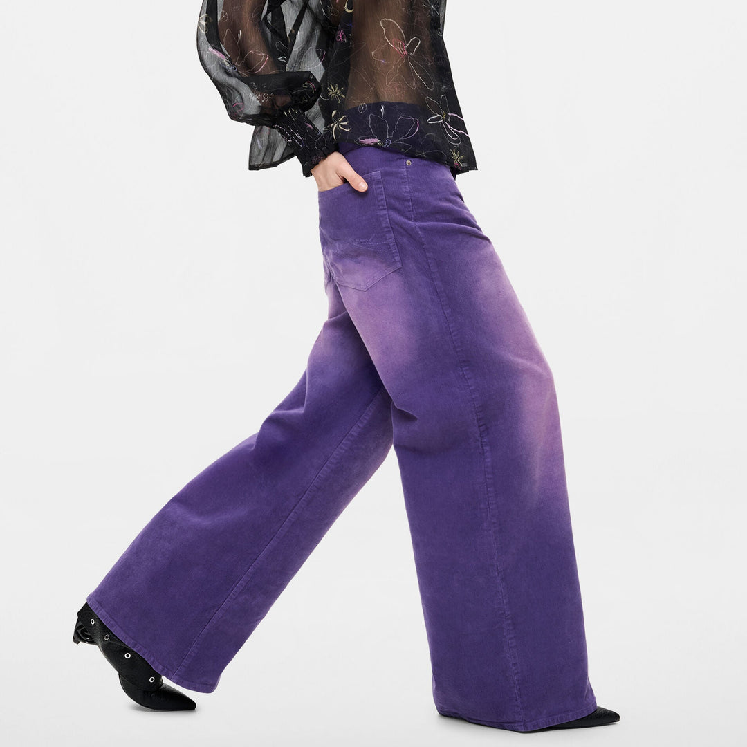 A woman modeling a sheer black long-sleeve top with a subtle floral pattern and wide-leg purple trousers, shown from a side profile against a light gray studio background.