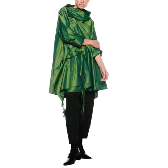 A model stands against a plain white studio background wearing a shiny metallic green poncho-style top with black pants and black pointed shoes.