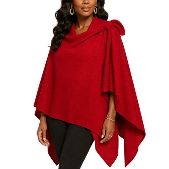 A woman modeling a vibrant red wrap poncho with a large bow detail on the shoulder, paired with black pants, against a plain light beige background.