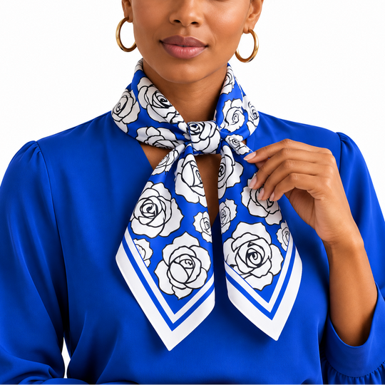 A woman wearing a royal blue blouse and a matching blue and white rose-patterned silk scarf, styled with gold hoop earrings against a solid white background.