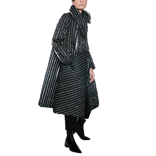 A model wearing a long black coat with thin white vertical stripes, paired with black pants and black ankle boots, photographed against a plain white studio background.
