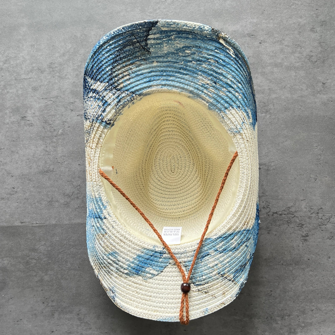 A woven straw hat viewed from above, featuring a cream-colored crown and brim with blue and white marbled or abstract painted patterns on the sides. It has an adjustable brown cord with a dark brown bead closure.
