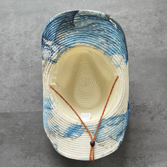 A woven straw hat viewed from above, featuring a cream-colored crown and brim with blue and white marbled or abstract painted patterns on the sides. It has an adjustable brown cord with a dark brown bead closure.