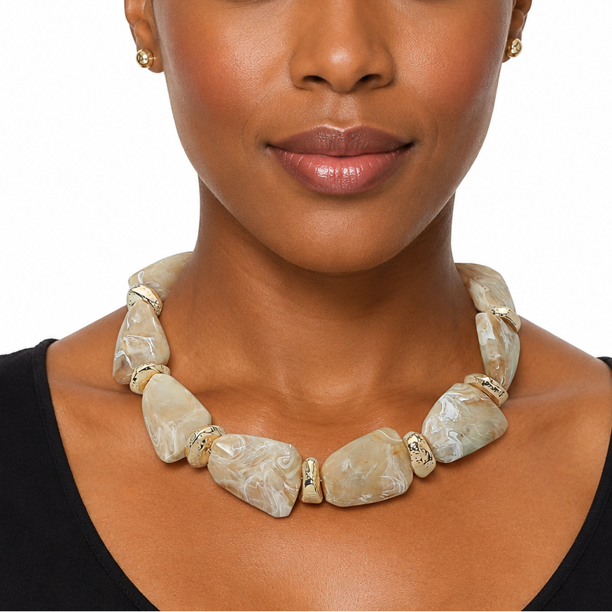 A woman wearing a chunky statement necklace made of irregular beige and cream stone-like beads with small gold spacer details, paired with matching small gold stud earrings, against a plain white background.