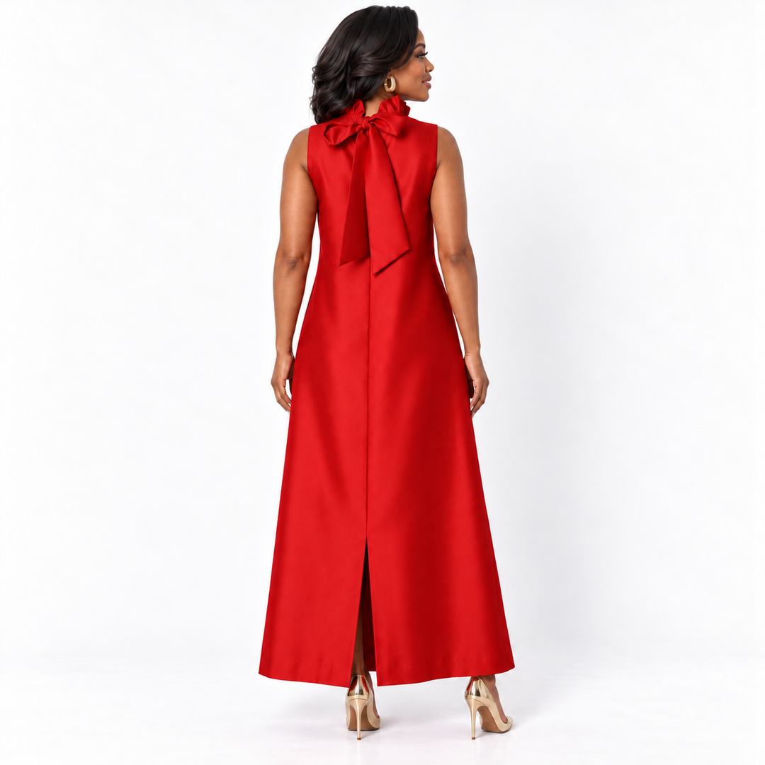 A woman modeling a sleeveless red maxi dress from the back, featuring a large bow at the back of the neck and a front slit, paired with gold high heels against a white studio background.