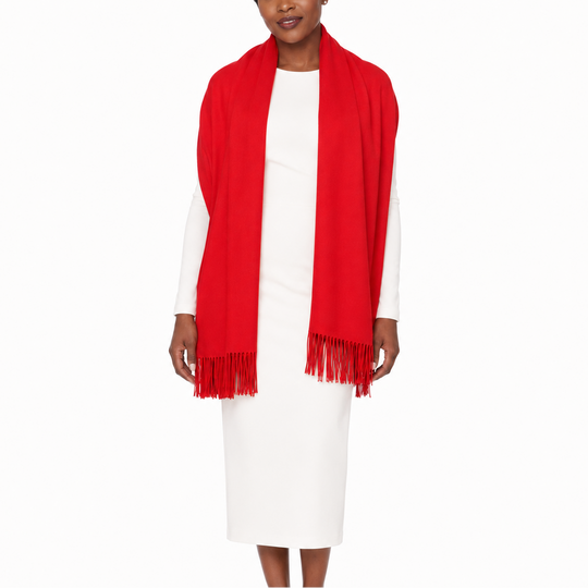 A woman modeling a bright red knit wrap or shawl with fringe trim, worn over a white long-sleeve dress against a plain white studio background.