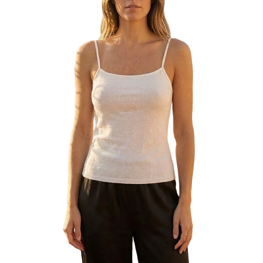 Woman wearing a white tank top and black pants on a rooftop with plants and furniture.