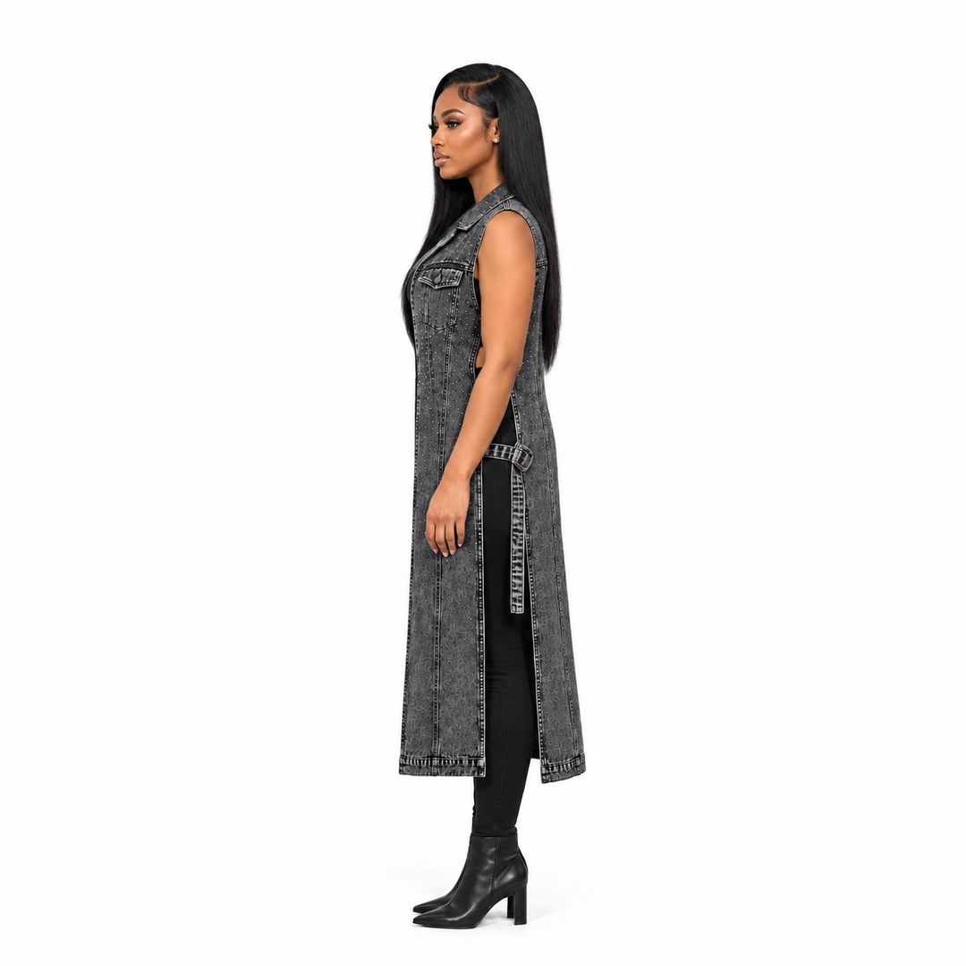 A woman shown in profile wearing a long sleeveless gray denim vest over a black outfit with black heeled ankle boots, photographed against a plain white studio background.