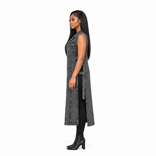 A woman shown in profile wearing a long sleeveless gray denim vest over a black outfit with black heeled ankle boots, photographed against a plain white studio background.