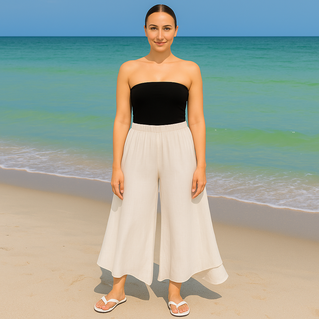 Woman in black strapless top and white pants standing on a beach with clear blue water and sky.