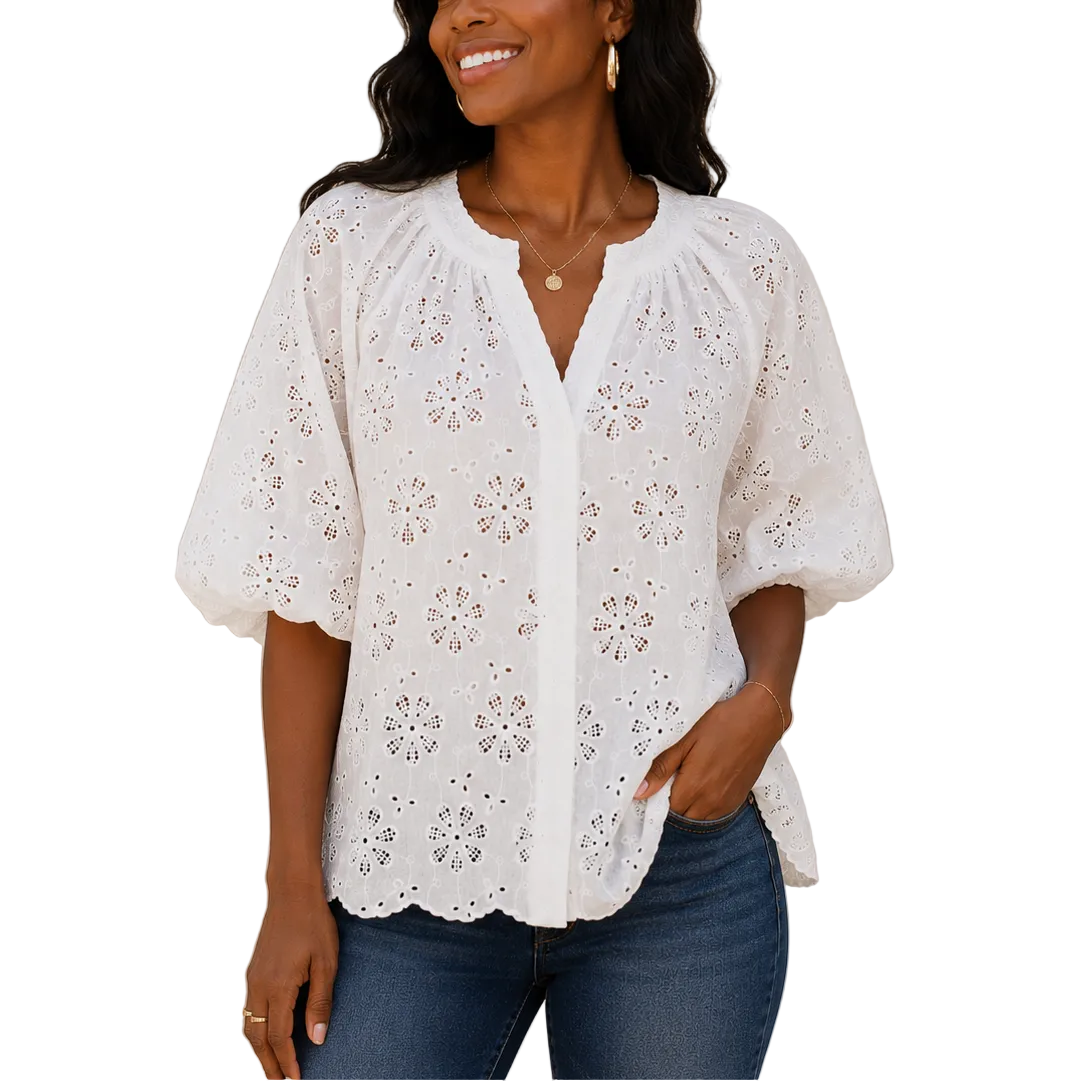 Woman wearing a white embroidered blouse and blue jeans standing against a neutral wall with a plant.