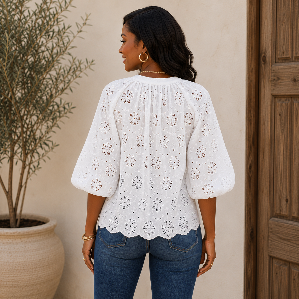 Woman wearing a white lace top and blue jeans standing against a beige wall with a wooden door.