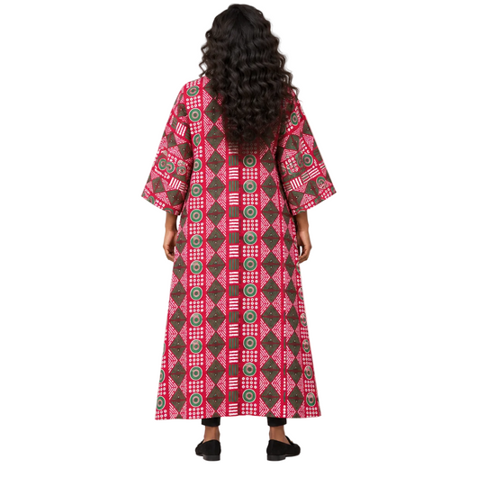 A woman viewed from behind wearing a long, wide-sleeved kaftan dress with a vibrant pink, brown, and green geometric pattern, set against a plain light gray studio background.