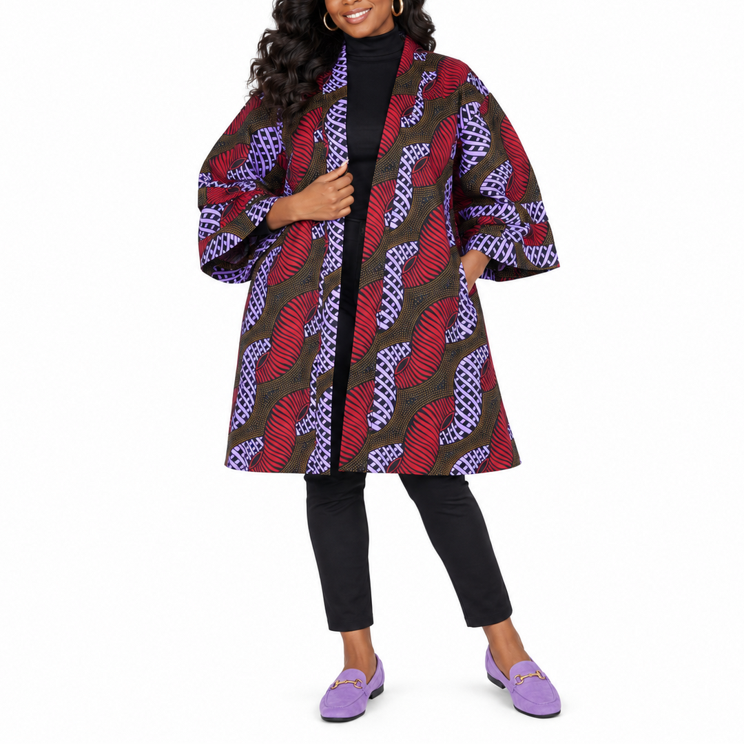 A woman modeling a long open-front coat with wide three-quarter sleeves, featuring a bold abstract pattern in red, purple, black, and brown over a black turtleneck, black pants, and purple loafers, set against a plain white background.