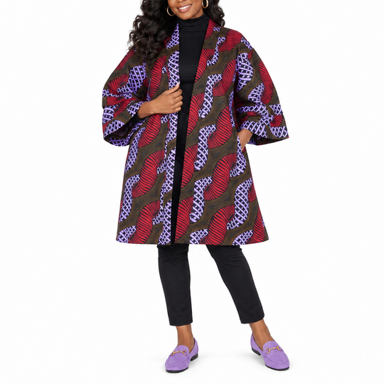 A woman modeling a long open-front coat with wide three-quarter sleeves, featuring a bold abstract pattern in red, purple, black, and brown over a black turtleneck, black pants, and purple loafers, set against a plain white background.