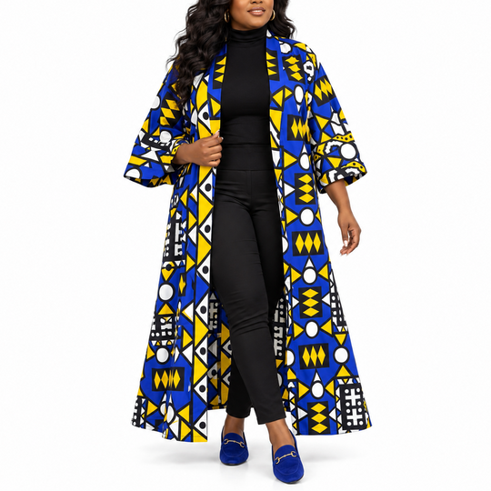 A woman modeling a long, open-front coat with a bold geometric African wax print in blue, yellow, black, and white, worn over a black outfit and blue loafers against a plain white background.