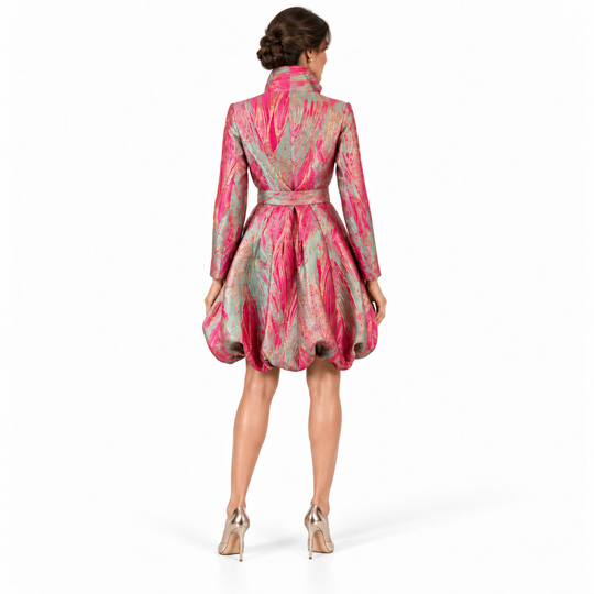 A woman viewed from the back wearing a short, structured jacket with a high collar, long sleeves, and a matching waist tie. The jacket features an abstract, painterly pattern in vibrant pink and shimmering metallic gold.