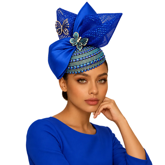A woman wearing an elaborate royal blue headwrap hat with sequins, beadwork, and butterfly embellishments, paired with a matching blue top, posed in a luxurious gold-toned boutique interior.