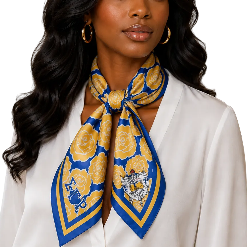 A woman with long, wavy black hair models a square silk scarf with a yellow and blue floral pattern, tied around her neck. She is wearing a white blouse and gold hoop earrings in an indoor setting with a neutral beige background.