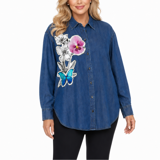 A woman modeling a blue denim button-up shirt with a large embroidered floral and butterfly design on the front, paired with black pants against a plain white background.