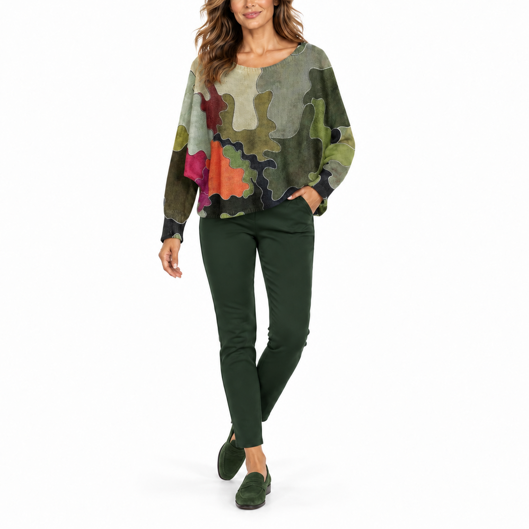 A woman modeling a long-sleeve bohemian-style top with a multicolor abstract print, paired with dark green pants and green suede loafers, against a solid white background.