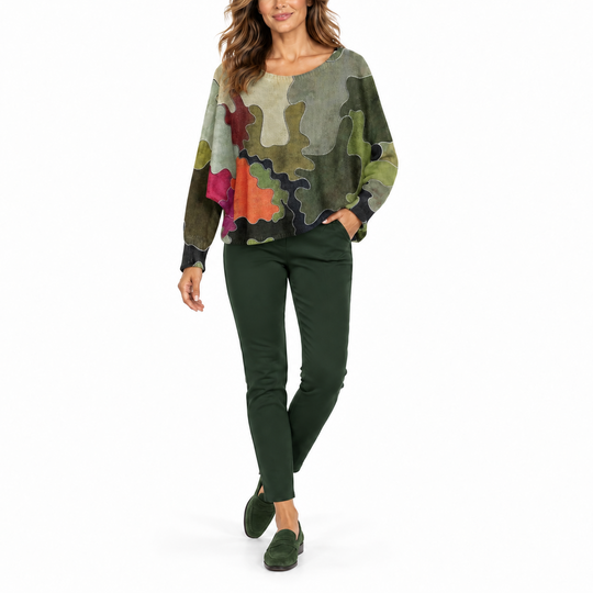 A woman modeling a long-sleeve bohemian-style top with a multicolor abstract print, paired with dark green pants and green suede loafers, against a solid white background.