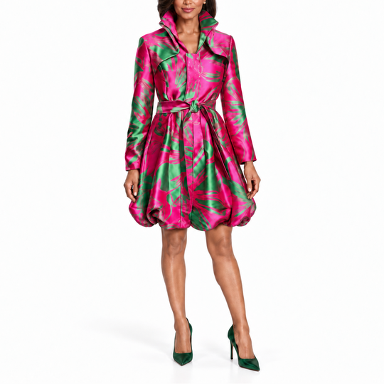 A woman modeling a short satin-like trench-style dress with a bold pink and green abstract print, matching green pointed heels, against a plain white studio background.
