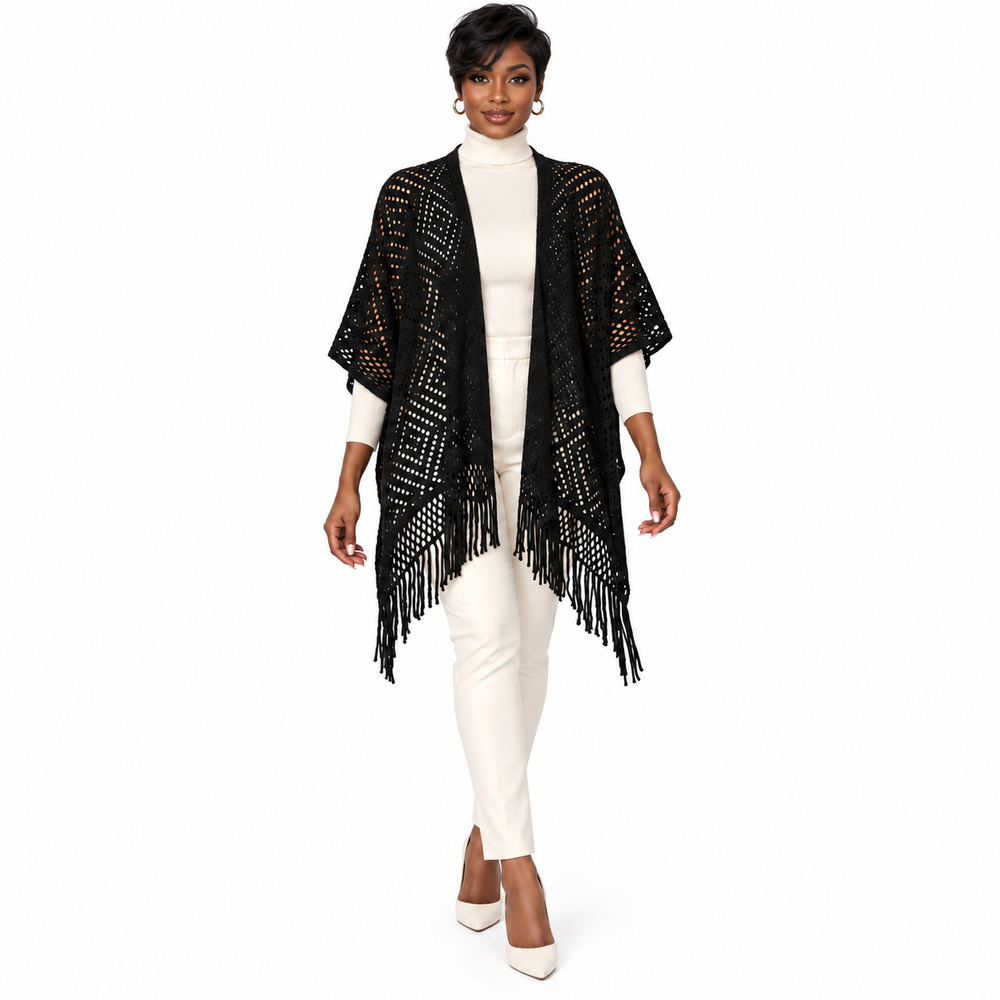 A woman modeling a black open-front knit poncho with fringe trim over a white turtleneck and white pants, photographed against a plain white studio background.