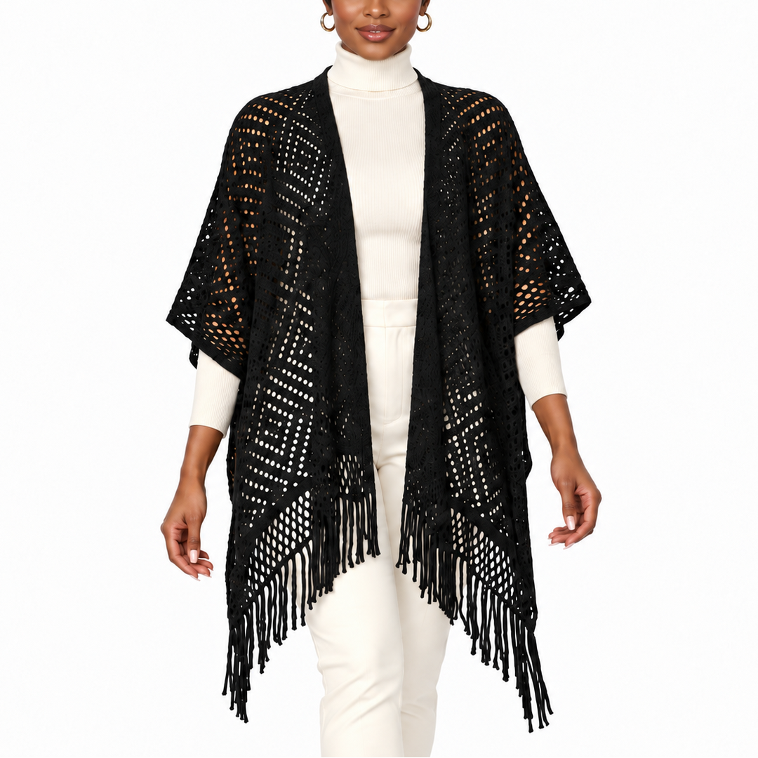 A woman modeling a black open-front fringed poncho-style cardigan over a white mock-neck top and white pants, photographed against a plain white studio background.
