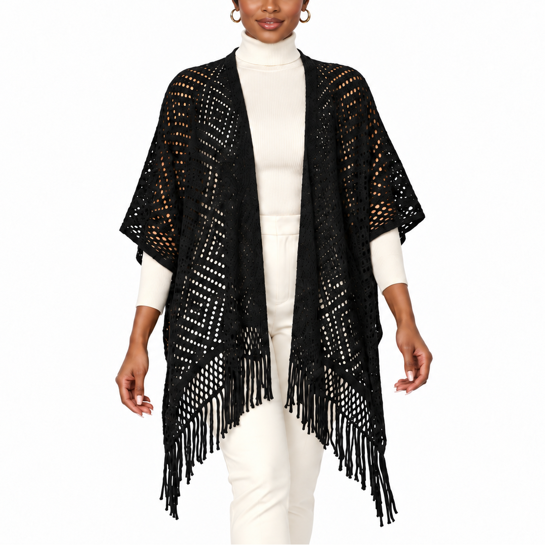 A woman modeling a black open-front fringed poncho-style cardigan over a white mock-neck top and white pants, photographed against a plain white studio background.