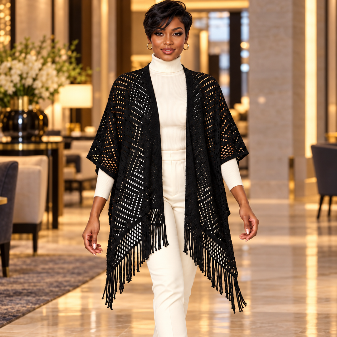 A woman walks forward in a luxury indoor hallway wearing a black open-front fringed poncho over a white turtleneck and white pants.