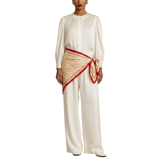 A model stands in a beige arched studio setting wearing a white long-sleeve blouse and matching wide-leg trousers, with a beige patterned scarf wrapped around the waist like a sash. A black box labeled SILKSILKY sits on the floor to the right.