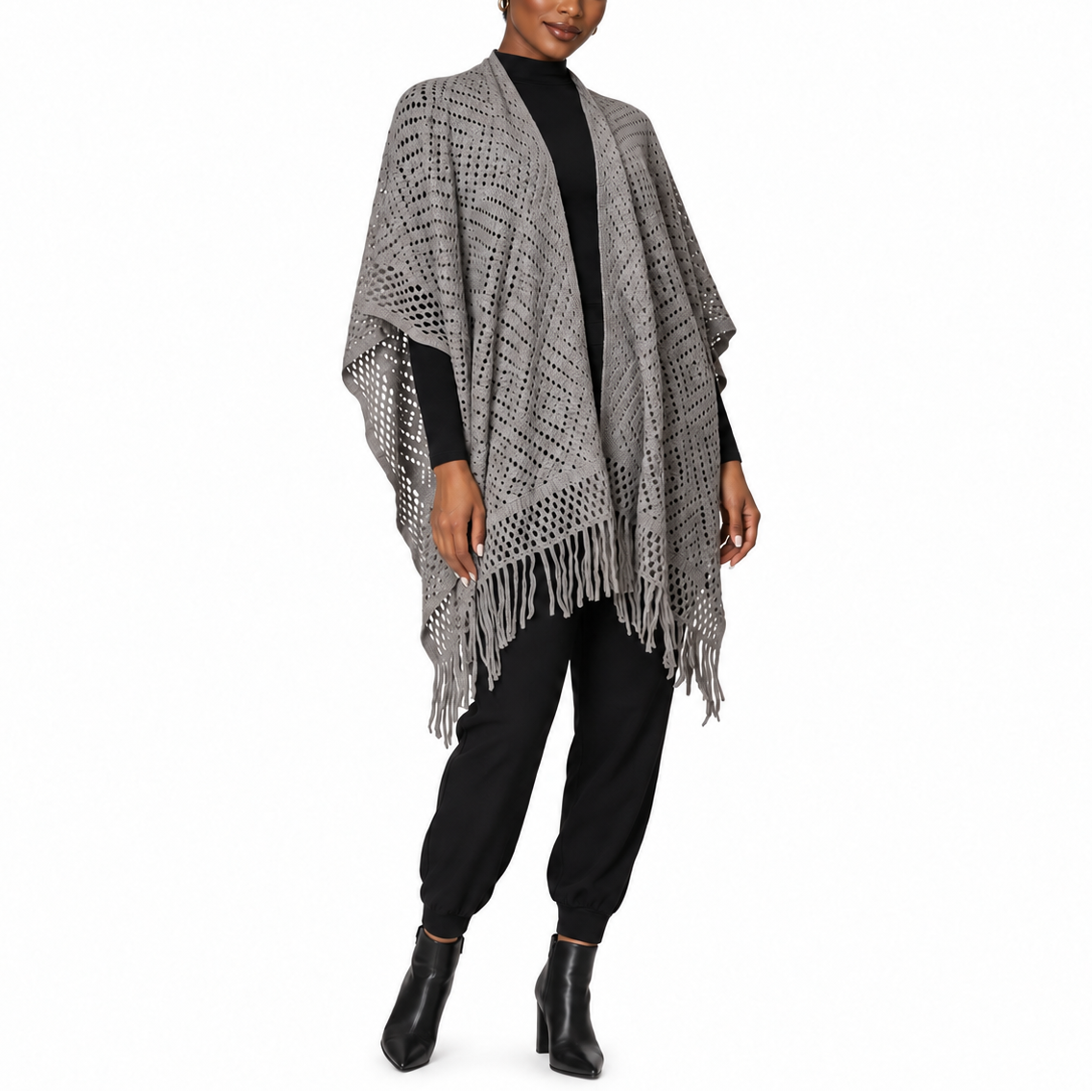 A woman modeling a gray open-knit poncho with fringe trim over a black long-sleeve top, black pants, and black ankle boots, photographed against a plain white studio background.