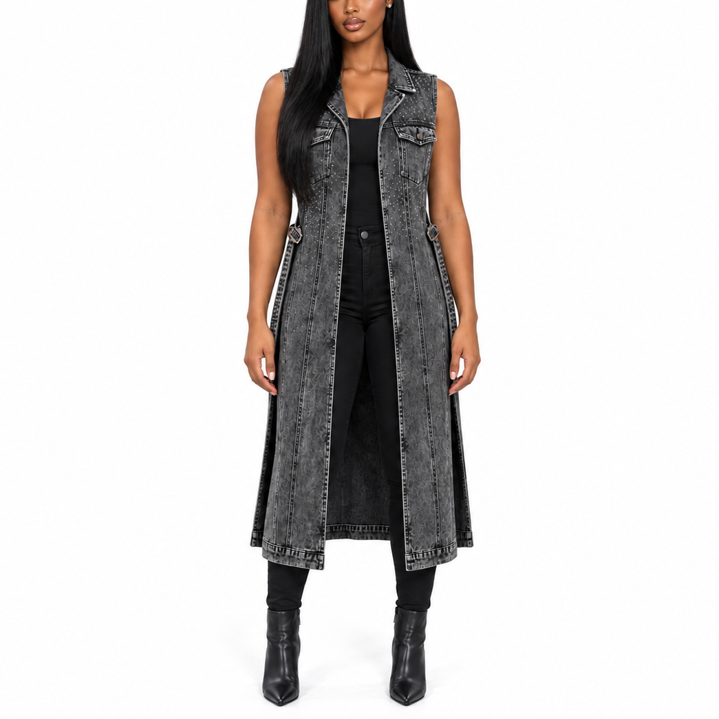 A woman modeling a long, sleeveless, charcoal gray washed denim vest with silver-tone studs and piping, worn over a black top and black pants, against a plain white background.