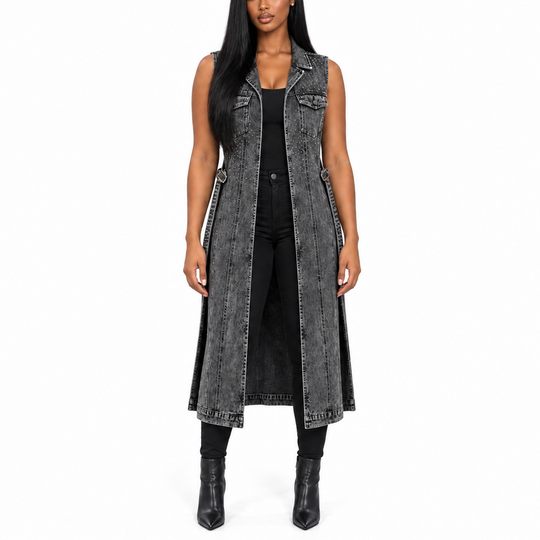 A woman modeling a long, sleeveless, charcoal gray washed denim vest with silver-tone studs and piping, worn over a black top and black pants, against a plain white background.