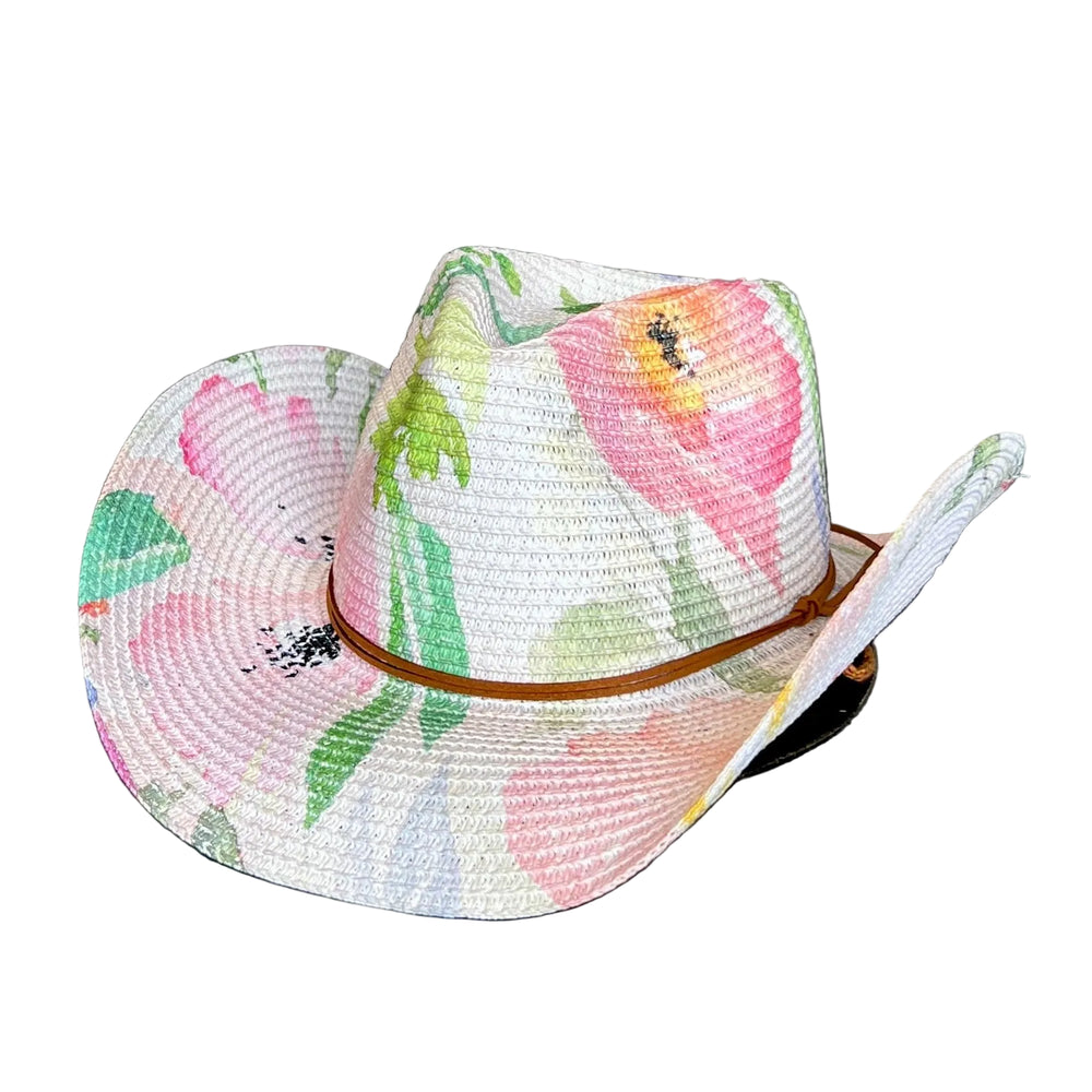 A woven straw cowboy hat with a colorful floral print in pink, green, and yellow, featuring a brown leather band, resting on a dark gray wooden surface against a gray concrete wall.