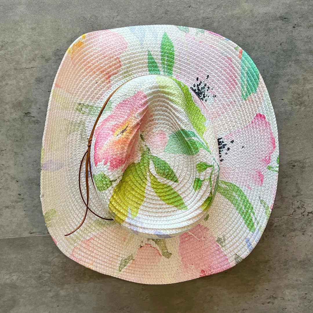A woven straw sun hat with a colorful floral print featuring pink flowers and green leaves on a light cream background, resting on a gray concrete surface.