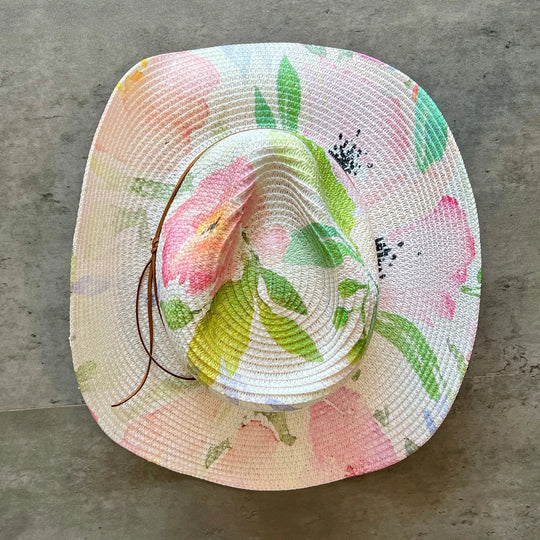 A woven straw sun hat with a colorful floral print featuring pink flowers and green leaves on a light cream background, resting on a gray concrete surface.