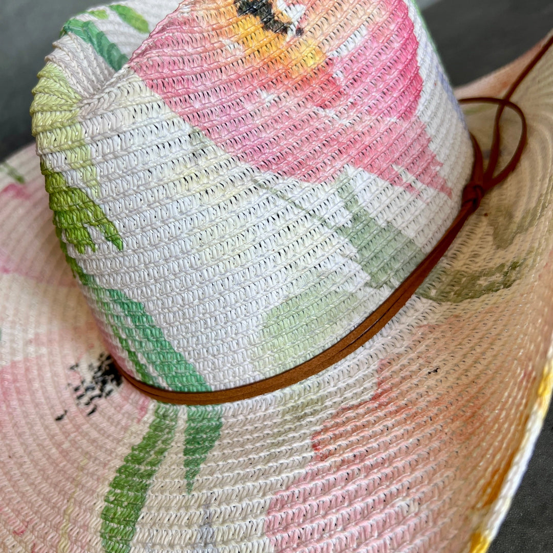 A close-up of a woven straw cowboy hat featuring a colorful floral print in pink, green, yellow, and orange, with a brown rope band.