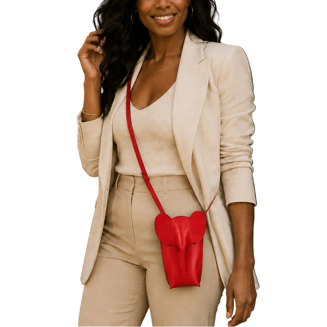A woman wearing a beige blazer and matching pants carries a small red heart-shaped crossbody bag with a red strap, photographed outdoors in front of a cafe setting.