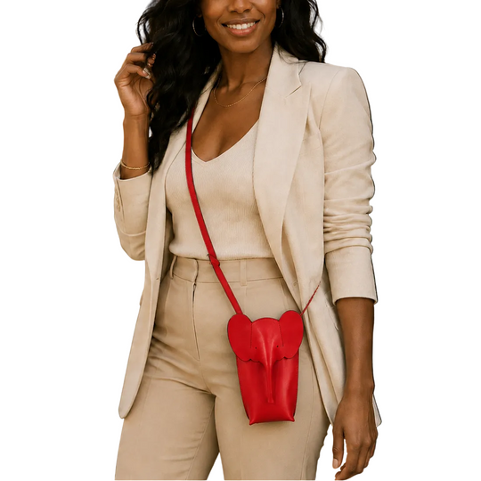 A woman wearing a beige blazer and matching pants carries a small red heart-shaped crossbody bag with a red strap, photographed outdoors in front of a cafe setting.